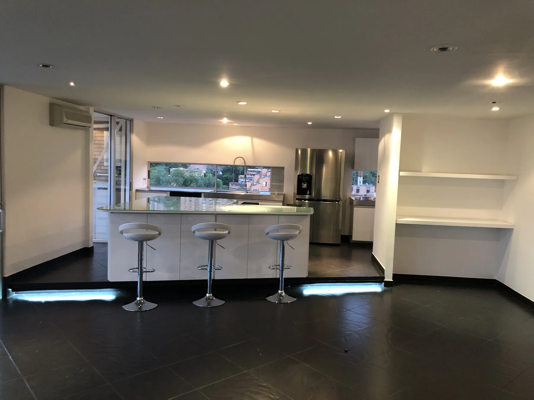Open-plan kitchen with white lacquer cabinetry, a backlit glass island counter and three chrome barstools