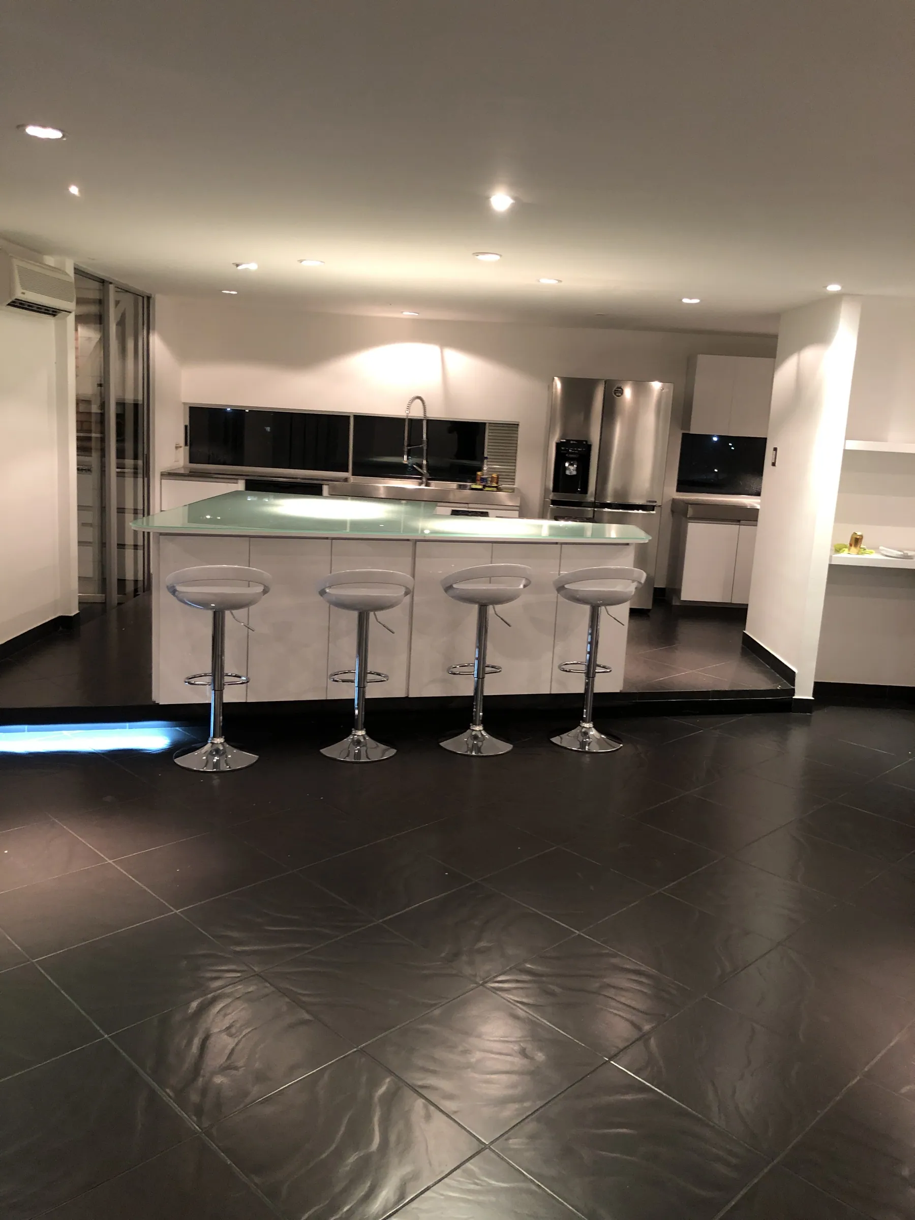 The kitchen at night, lit island counter with four barstools and stainless steel appliances reflected in polished stone floors